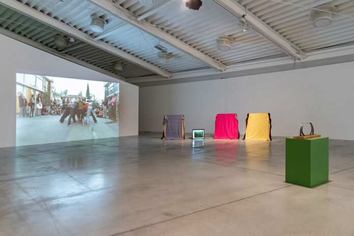 Simone Forti at Pecci center la foto a colori mostra due poltrone ricoperte rispettivamente da un telo giallo e un telo fucsia, un'opera d'arte simile ad un anello posta su un piedistallo verde chiaro e uno schermo che riproduce un'immagine di gente in lotta come nel rugby