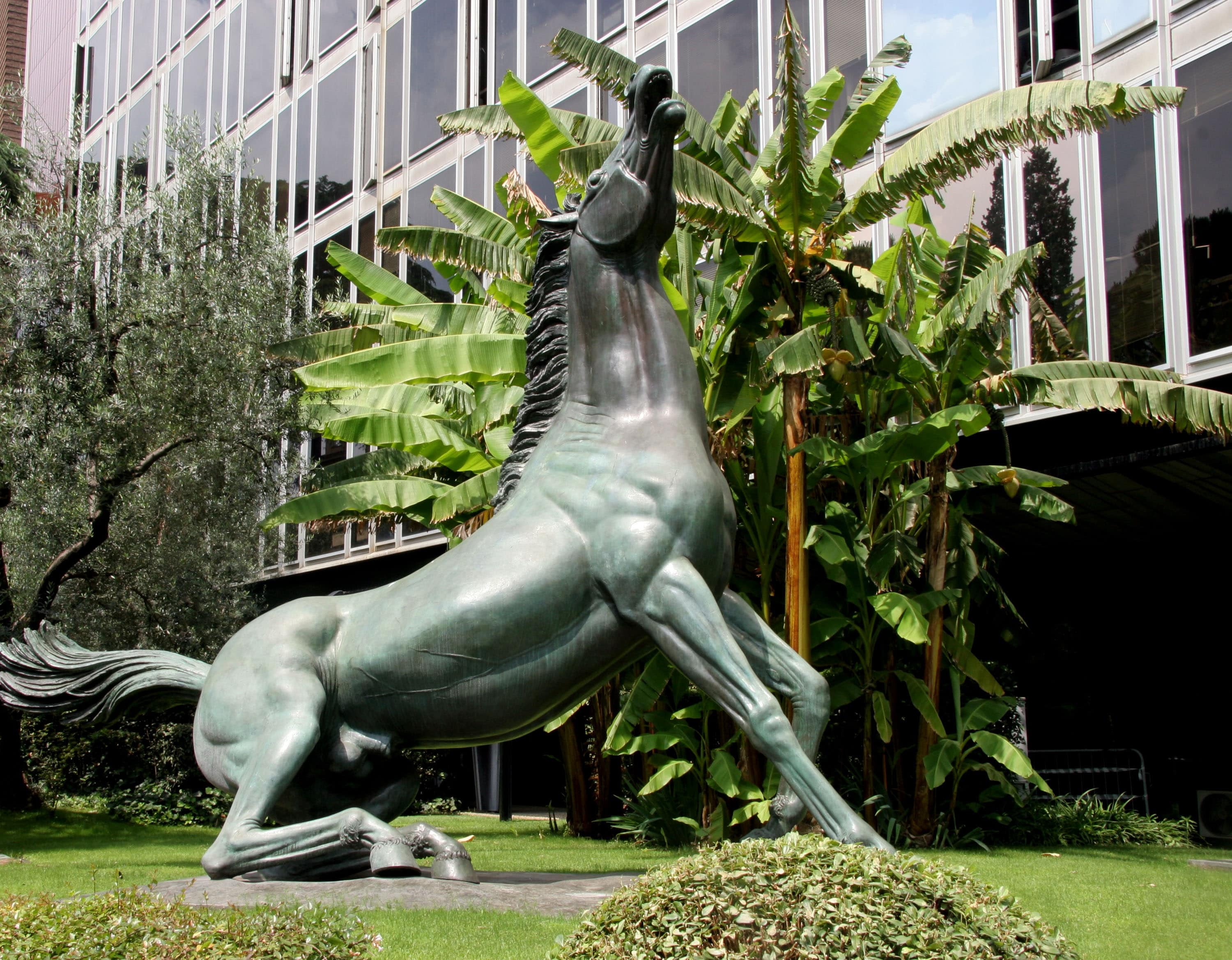 scultura raffigurante un cavallo che nitrisce sollevando il muso verso l'alto. La scultura è all'interno di uno spazio verde, davanti alle vetrate di un edificio