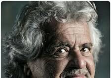 L'immagine mostra l'ex-comico e fondatore del Movimento 5 Stelle Beppe Grillo. Un uomo dai capelli bianchi e mossi sebbene corti con un accenno di barba bianca. La foto è a colori desaturati