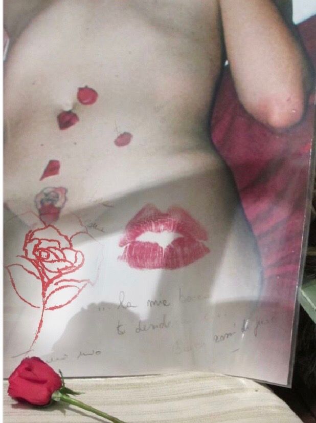 la foto a colori mostra una rosa rossa con gambo e l'immagine di parte di un corpo di donna dipinto con labbra rosse e dedica