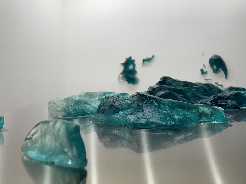 L'immagine mostra un particolare dell’opera di Margherita Moscardini presso la galleria Renata Fabbri. L'opera è una massa vetrosa informe dalle tonalità verde acqua più o meno luminose