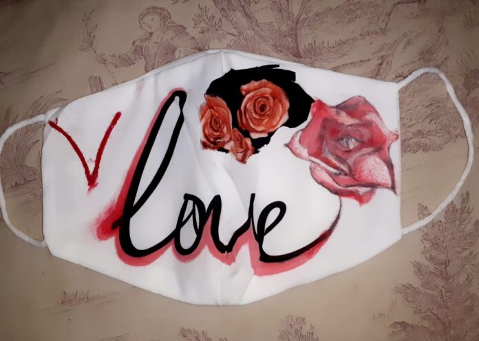 immagine di una mascherina bianca con la scritta love in rosso e nero. In alto e a destra della scritta ci sono delle rose, una più grande e tre più piccole su sfondo nero