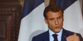 L'immagine mostra il presidente francese Emmanuel Macron durante un discorso. Il politico ha corti capelli bruni, indossa una giacca blu su camicia bianca e cravatta blu nera e sullo sfondo sono visibili il pezzo di una porta in legno e due bandiere francesi delle quali si vedono solo il bianco e il blu