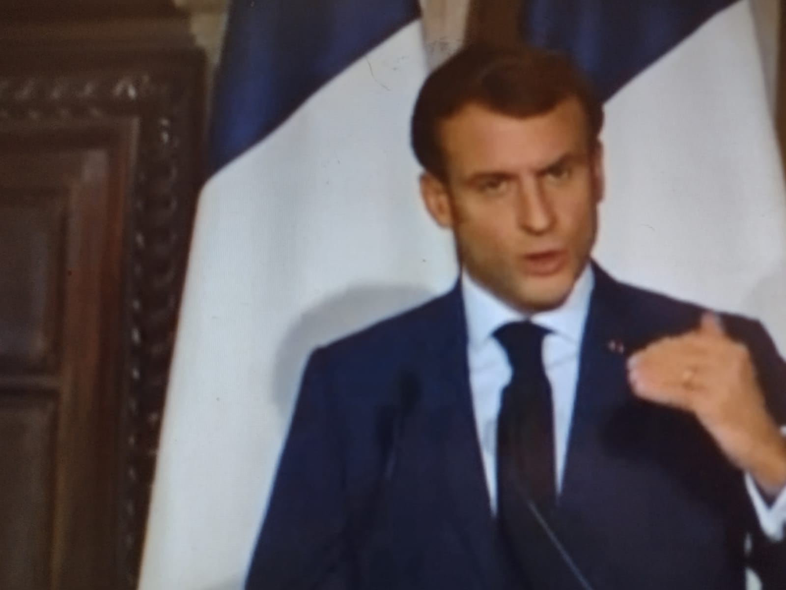 L'immagine mostra il presidente francese Emmanuel Macron durante un discorso. Il politico ha corti capelli bruni, indossa una giacca blu su camicia bianca e cravatta blu nera e sullo sfondo sono visibili il pezzo di una porta in legno e due bandiere francesi delle quali si vedono solo il bianco e il blu