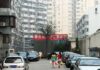L'immagine mostra una via della capitale Cinese, immediatamente riconoscibile per la scritta in ideogrammi bianchi su fondo rosso al centro di una distresa di palazzi grigi con un piccolo alberello nel mezzo. In basso è visibile una strada con i bordi occupati da macchina parcheggiate e due donne che portano la spesa verso i palazzi