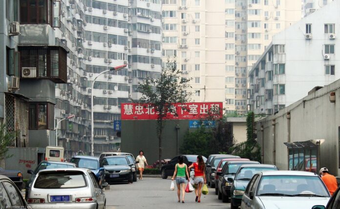 L'immagine mostra una via della capitale Cinese, immediatamente riconoscibile per la scritta in ideogrammi bianchi su fondo rosso al centro di una distresa di palazzi grigi con un piccolo alberello nel mezzo. In basso è visibile una strada con i bordi occupati da macchina parcheggiate e due donne che portano la spesa verso i palazzi