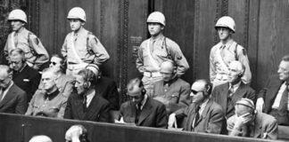 L'immagine è una foto in bianco e nero che mostra alcuni degli imputati al processo di Norimberga nel 1945. Gli imputati siedono dietro dei banchi con cuffie mentre ascoltano le accuse sotto l'occhio vigile di guardie riconoscibili dalla divisa