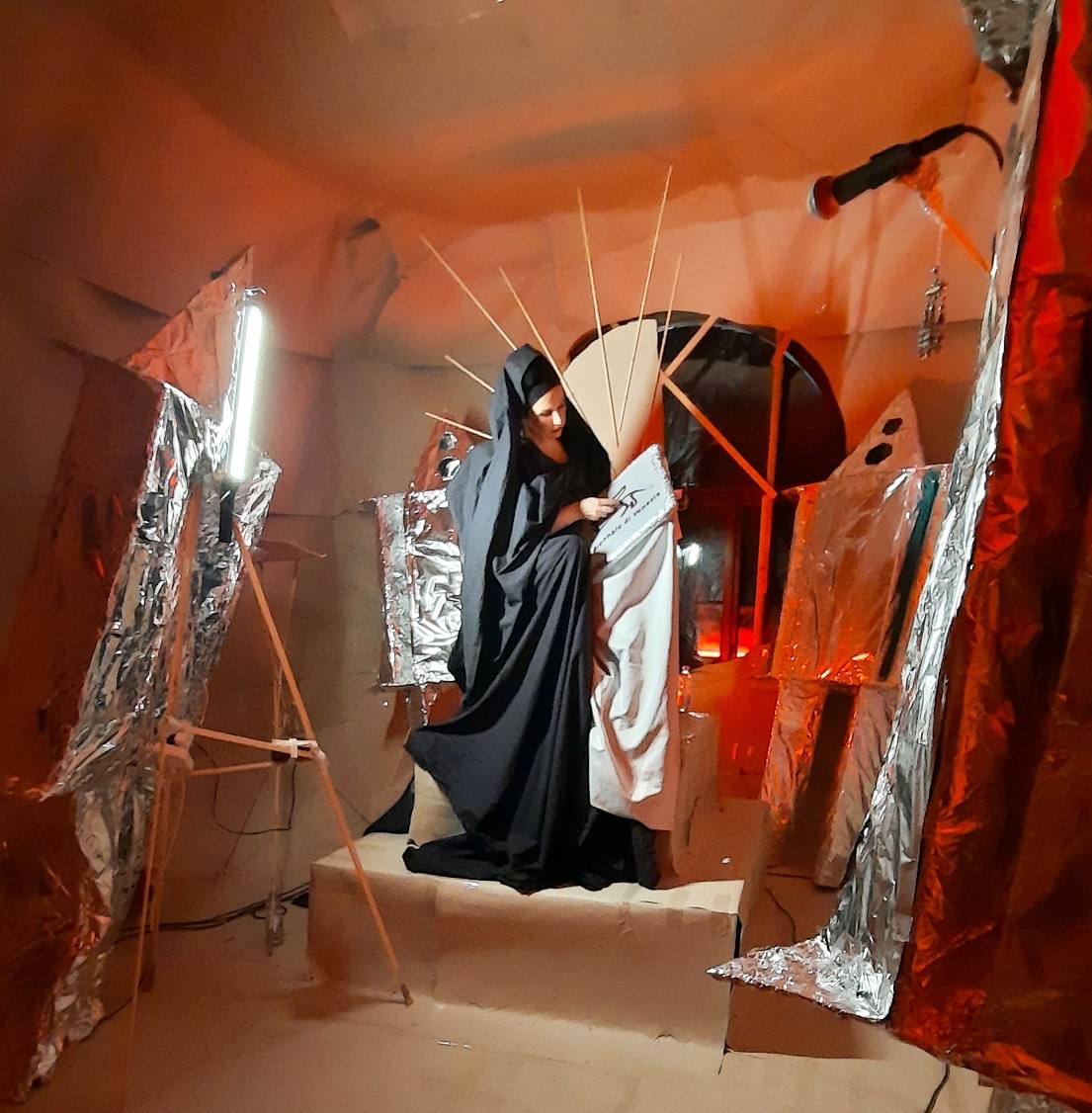 L'immagine mostra un'opera di una degli artisti del collettivo ORTA in mostra alla Biennale di Venezia del 2022. L'opera è un'installazione consistente in una donna vestita interamente di nero intenta a tessere al centro di una camera coperta di tessuto e circondata da cavalletti coperti di carta argentata