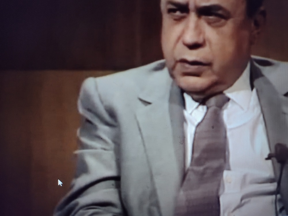 L'immagine mostra lo scrittore Leonardo Sciascia in una ripresa televisiva. Lo scrittore siciliano occupa la parte destra dell'immagine. Indossa giacca e cravatta grigie su camicia bianca. Il fondo è marrone